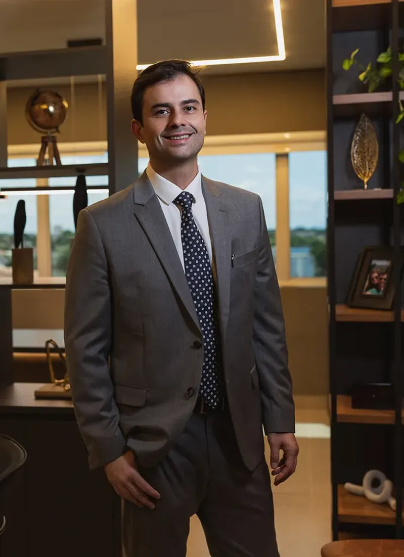 Rodrigo Marçal, advogado sorridente com terno cinza e gravata azul de bolinhas, posando em escritório sofisticado com estantes decoradas, globo dourado, iluminação quente e vista para área verde ao fundo, fotografia profissional realista com atmosfera elegante e acolhedora